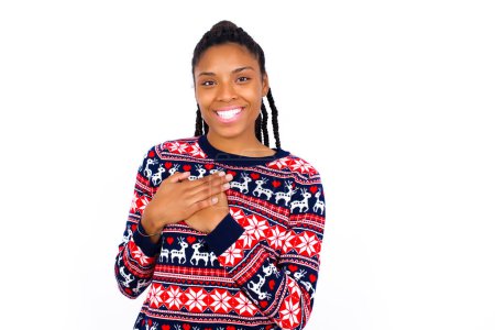 Foto de Honesta mujer afroamericana vistiendo suéter de Navidad contra la pared blanca mantiene las manos en el pecho, tocado por el cumplido o hace la promesa, mira a la cámara con gran placer. - Imagen libre de derechos