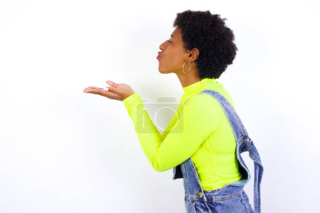 Téléchargez les photos : Profil vue de côté portrait de la jeune femme afro-américaine attrayante avec les cheveux courts portant le denim ensemble contre le mur blanc envoyant baiser d'air - en image libre de droit