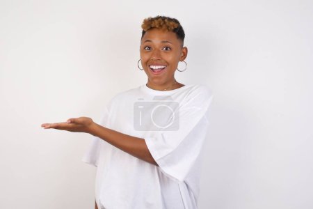 Foto de La joven afro-americana alegre positiva que usa la camiseta blanca dice: wow qué emocionante es, tiene expresión sorprendida, muestra algo en el espacio en blanco con la mano abierta sobre el fondo gris aislado. Concepto de publicidad. - Imagen libre de derechos