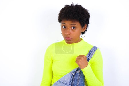 Photo for Young African American woman with short hair wearing denim overall against white wall shows fist has annoyed face expression going to revenge or threaten someone makes serious look. I will show you who is boss - Royalty Free Image