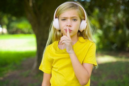 Photo for Caucasian little girl wearing yellow t-shirt standing outdoors making hush gesture with finger on her lips wearing  wireless headphones. Be quiet. - Royalty Free Image
