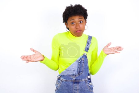 Photo for Puzzled and clueless young African American woman with short hair wearing denim overall against white with arms out, shrugging shoulders, saying: who cares, so what, I don't know. Negative human emotions. - Royalty Free Image