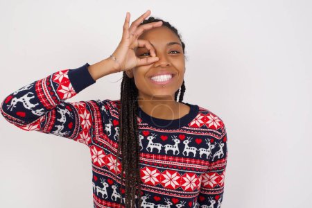 Téléchargez les photos : Femme afro-américaine portant un pull de Noël contre un mur blanc avec un visage heureux souriant faisant signe ok avec la main sur les yeux en regardant à travers le doigt. - en image libre de droit