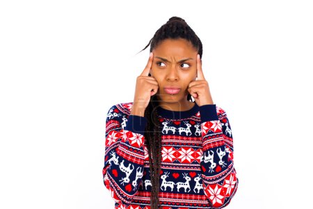 Téléchargez les photos : Femme afro-américaine portant un pull de Noël contre un mur blanc avec une expression réfléchie, regarde ailleurs, garde la main près du visage, pense à quelque chose d'agréable. - en image libre de droit