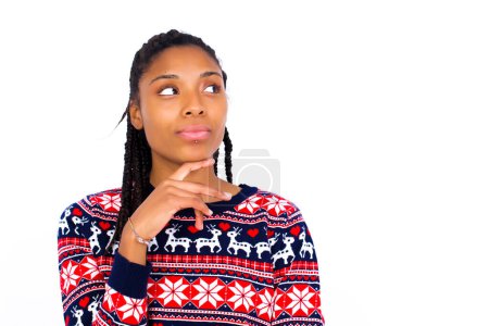 Photo for Dreamy African American woman wearing Christmas sweater against white wall with pleasant expression, looks sideways, keeps hand under chin, thinks about something pleasant. - Royalty Free Image
