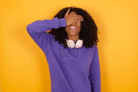 Téléchargez les photos : Portrait de jeune femme afro-américaine expressive avec écouteurs sur fond jaune couvrant les yeux avec la main - en image libre de droit
