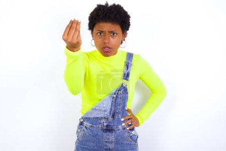 Foto de What the hell are you talking about. Shot of frustrated young African American woman with short hair wearing denim overall against white gesturing with raised hand doing Italian gesture, frowning, being displeased and confused with dumb question. - Imagen libre de derechos