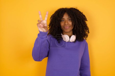 Photo for Portrait of young expressive african american woman with headphones on yellow background showing v sign - Royalty Free Image