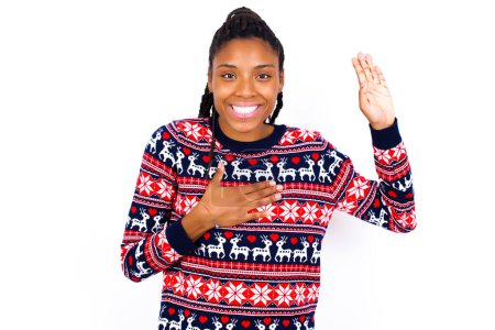 Foto de Lo juro, prométeme que no te arrepentirás. Retrato de una mujer afroamericana sincera usando suéter de Navidad contra la pared blanca levantando un brazo y tomándose de la mano en el corazón como dar juramento, diciendo la verdad, quiero que creas. - Imagen libre de derechos
