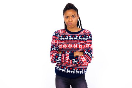 Foto de Imagen de una mujer afroamericana enojada usando suéter navideño contra una pared blanca cruzando brazos. Mirando a la cámara con expresión decepcionada. - Imagen libre de derechos