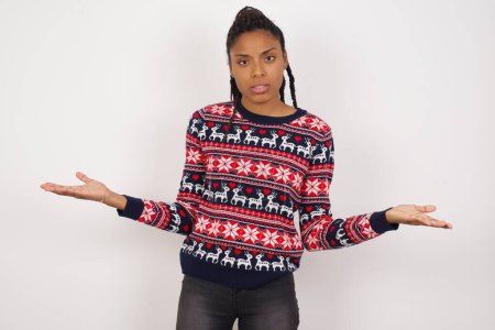 Téléchargez les photos : Femme afro-américaine portant un pull de Noël contre un mur blanc semble épaules hausseurs d'épaules incertaines. - en image libre de droit