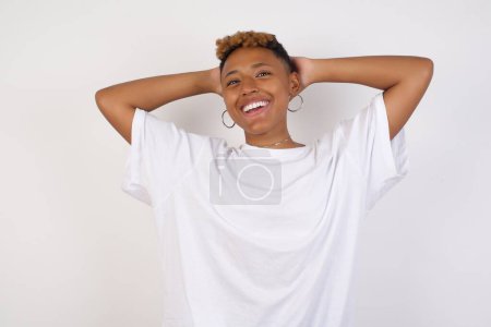 Téléchargez les photos : Concept de confiance et de coquetterie. Portrait de charmante jeune femme afro-américaine portant un t-shirt blanc souriant largement avec une expression assurée tout en se tenant la main sur la tête. Se placer dans le contexte. - en image libre de droit