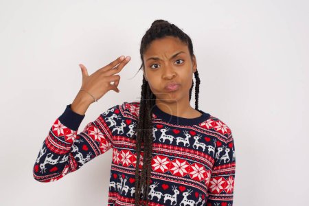 Foto de Mujer afroamericana infeliz vistiendo suéter de Navidad contra pared blanca imita disparo de arma hace gesto suicida mantiene dos dedos en las sienes. - Imagen libre de derechos