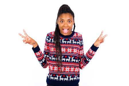 Téléchargez les photos : Jeune belle femme afro-américaine portant un pull de Noël contre un mur blanc avec un sourire optimiste, montrant un geste de paix ou de victoire avec les deux mains, l'air amical. Signe V. - en image libre de droit