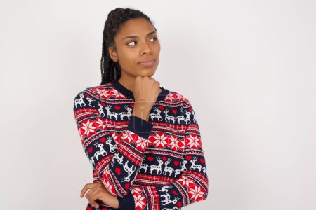 Téléchargez les photos : Femme afro-américaine portant un pull de Noël contre un mur blanc avec la main sous le menton et regardant de côté avec une expression douteuse et sceptique, suspecte et douteuse. - en image libre de droit