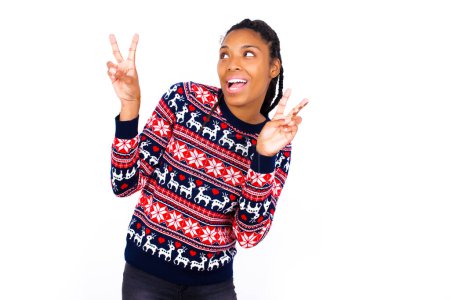Photo for Isolated shot of cheerful African American woman wearing Christmas sweater against white wall makes peace or victory sign with both hands, feels cool. - Royalty Free Image
