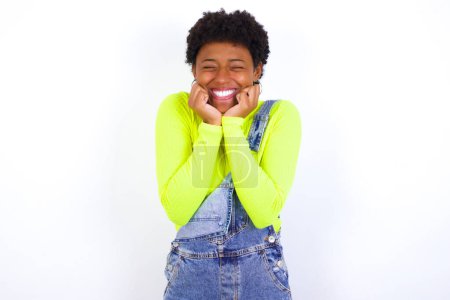 Foto de Retrato de una joven afroamericana con el pelo corto usando denim en general contra la pared blanca siendo abrumado, expresando emoción y felicidad con los ojos cerrados y las manos cerca de la cara. - Imagen libre de derechos