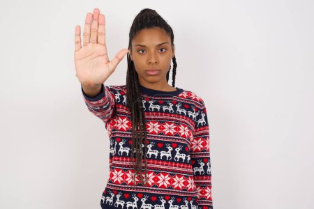 Foto de Mujer afroamericana vistiendo jersey navideño contra pared blanca haciendo gesto de stop con palma de la mano. Expresión de advertencia con gesto negativo y serio en la cara. - Imagen libre de derechos
