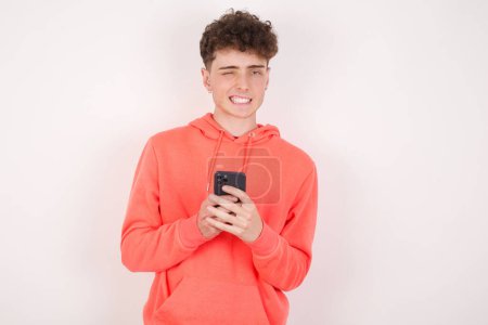 Téléchargez les photos : Beau jeune homme agréable en utilisant l'auto-téléphone et en regardant et en clignant des yeux à la caméra. Concept flirt et coquette. - en image libre de droit