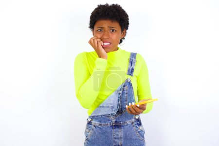 Foto de Miedo divertida mujer afroamericana joven con el pelo corto usando denim en general contra la pared blanca sosteniendo el teléfono y mordiendo clavos - Imagen libre de derechos