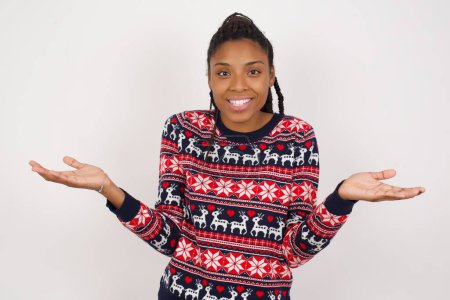 Photo for So what? Portrait of arrogant young beautiful African American woman wearing Christmas sweater against white wall shrugging hands sideways smiling gasping indifferent, telling something obvious. - Royalty Free Image