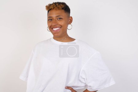 Téléchargez les photos : Photo studio d'une jeune afro-américaine joyeuse portant un t-shirt blanc garde la main sur la hanche, sourit largement, anticipe pour la surprise préparée par des amis le jour de son anniversaire, isolée sur fond. - en image libre de droit