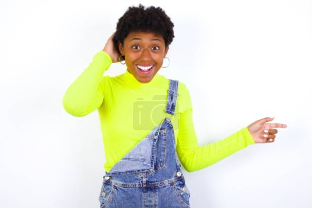 Foto de Mujer afroamericana joven sorprendida con el pelo corto usando denim en general contra la pared blanca apuntando al espacio vacío sosteniendo la mano en la cabeza - Imagen libre de derechos