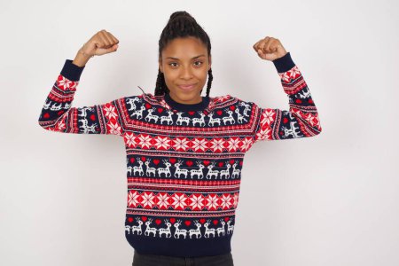 Téléchargez les photos : Tour de taille de femme afro-américaine portant un pull de Noël contre un mur blanc lève les bras pour montrer les muscles se sent confiant dans la victoire, semble forte et indépendante, sourit positivement à la caméra. Concept sportif. - en image libre de droit