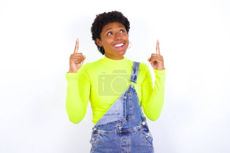 Foto de Exitosa mujer afroamericana de aspecto amigable con pelo corto que usa denim en general contra la pared blanca exclamando con entusiasmo, apuntando con ambos dedos índice hacia arriba, indicando algo. - Imagen libre de derechos