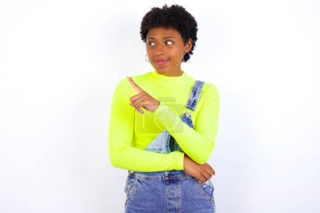 Foto de Retrato de una joven afroamericana con el pelo corto usando denim en general contra la pared blanca posando en la cámara con aspecto complicado, presentando el producto con el dedo índice. Concepto de publicidad. - Imagen libre de derechos