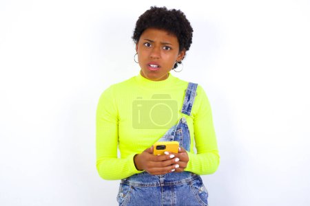 Photo for Photo of astonished crazy African American female with curly bushy wears jeans overalls over white wall hold smartphone dislike feedback concept - Royalty Free Image