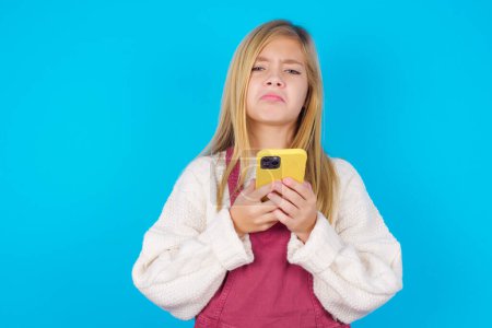 Photo for Portrait of a confused pretty girl holding mobile phone and shrugging shoulders and frowning face. - Royalty Free Image