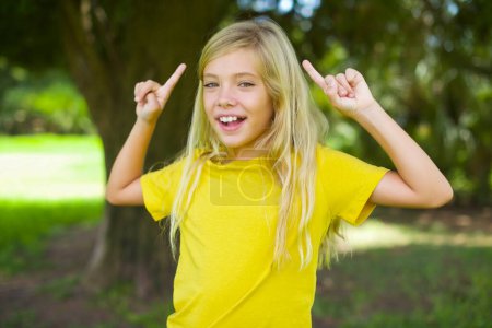 Téléchargez les photos : Joyeux caucasien petite fille portant jaune t-shirt debout à l'extérieur démontrant coiffure - en image libre de droit