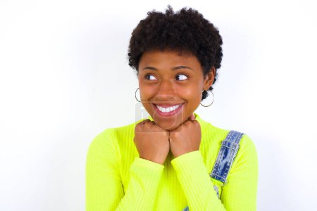 Téléchargez les photos : Jeune femme afro-américaine avec les cheveux courts portant le denim dans l'ensemble contre le mur blanc tient les mains sous le menton, heureux d'entendre des mots chaleureux de l'étranger - en image libre de droit