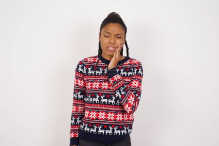 Téléchargez les photos : Concept de mal de dents. Plan intérieur de jeune femme afro-américaine portant un pull de Noël contre la douleur du mur blanc, tenant sa joue avec la main, souffrant de mal de dents, regardant la caméra avec une expression douloureuse - en image libre de droit