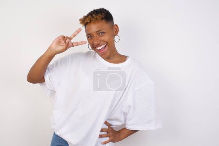 Photo for Leisure lifestyle people person celebrate flirt coquettish concept. Beautiful pretty young African American woman wearing white t-shirt showing v-sign near eyes wearing casual clothes standing indoors. - Royalty Free Image
