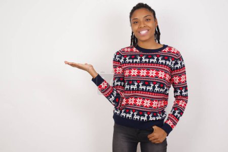 Foto de Positivo contenta mujer afroamericana vistiendo suéter de Navidad contra la pared blanca dice: wow lo emocionante que es, tiene expresión asombrada, muestra algo en el espacio en blanco con la mano abierta. Concepto de publicidad. - Imagen libre de derechos