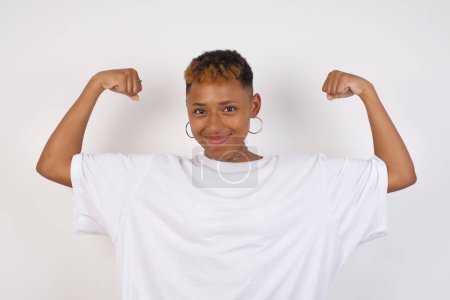 Photo for Waist up shot of african-american woman wearing white t-shirt raises arms to show her muscles feels confident in victory, looks strong and independent, smiles positively at camera, stands against gray background. Sport concept. - Royalty Free Image