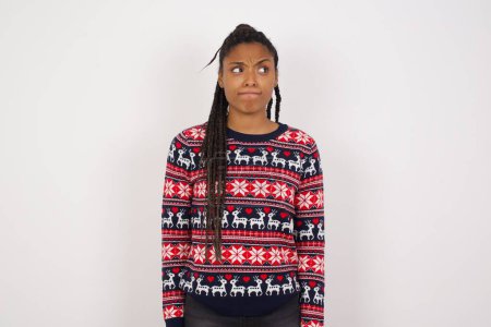 Foto de Sorprendido desconcertado joven hermosa mujer afroamericana con suéter de Navidad contra la pared blanca curva los labios y tiene mirada preocupada, ve algo horrible en frente. - Imagen libre de derechos