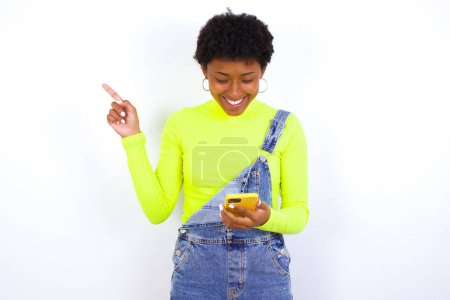 Photo for Young African American woman with short hair wearing denim overall against white wall using phone and pointing finger - Royalty Free Image