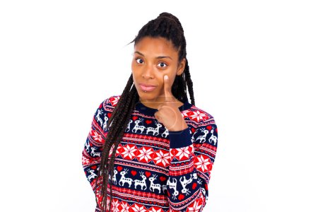Téléchargez les photos : Femme afro-américaine portant un pull de Noël contre un mur blanc regardant, observant, gardant un œil sur un objet devant, ou veillant sur quelque chose. - en image libre de droit