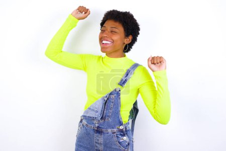 Foto de Foto de la joven mujer afroamericana optimista con el pelo corto con denim en general contra la pared blanca tiene diversión y baila despreocupado desgaste estar en perfecto estado de ánimo hace movimientos. Pasa tiempo libre en la fiesta disco - Imagen libre de derechos