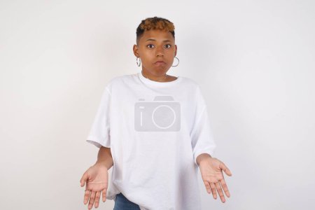 Photo for Puzzled and clueless young African American woman wearing white t-shirt with arms out, shrugging her shoulders, saying: who cares, so what, I don't know. Negative human emotions, facial expressions, life perception and attitude. - Royalty Free Image