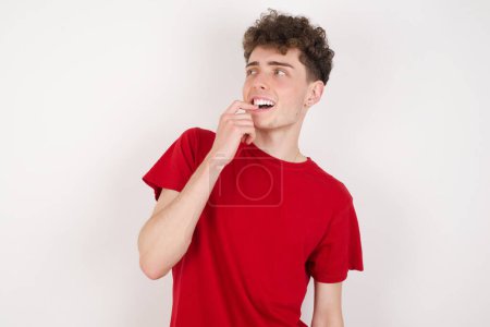 Photo for Young man with thoughtful expression, looks to the camera, keeps hand near face, bitting a finger thinks about something pleasant. - Royalty Free Image
