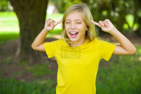 Foto de Foto de una chiquilla caucásica loca usando una camiseta amarilla al aire libre gritando y señalando con los dedos a los ojos cerrados del pelo - Imagen libre de derechos
