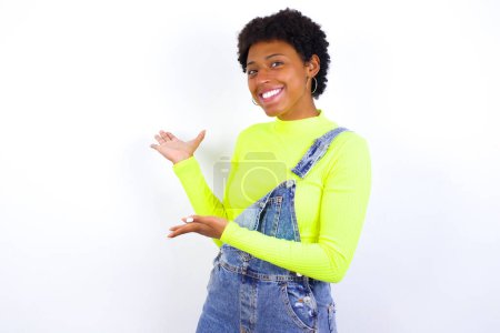 Foto de Joven mujer afroamericana con el pelo corto con denim en general contra la pared blanca Invitar a entrar sonriendo natural con las manos abiertas. Señal de bienvenida. - Imagen libre de derechos
