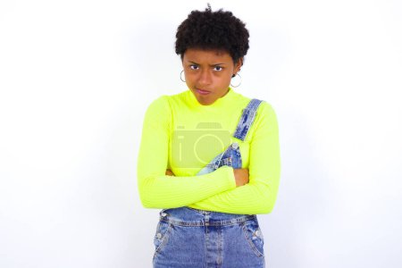 Téléchargez les photos : Photo de jeune femme afro-américaine en colère avec les cheveux courts portant le denim dans l'ensemble contre le mur blanc traversant les bras. Regardant la caméra avec une expression déçue. - en image libre de droit