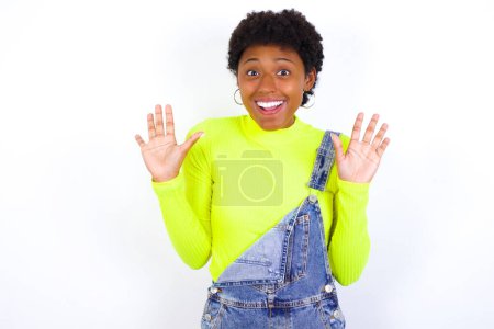 Photo for Delighted positive young African American woman with short hair wearing denim overall against white wall opens mouth  and arms palms up after having great result - Royalty Free Image