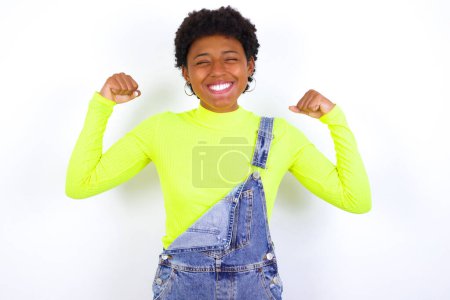 Téléchargez les photos : Forte puissante jeune femme afro-américaine aux cheveux courts portant le denim dans l'ensemble contre le mur blanc dents sourire, lève les bras et montre biceps. Regarde mes muscles.! - en image libre de droit