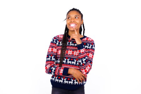 Photo for African American woman wearing Christmas sweater against white wall with thoughtful expression, looks to the camera, keeps hand near face, biting a finger thinks about something pleasant. - Royalty Free Image